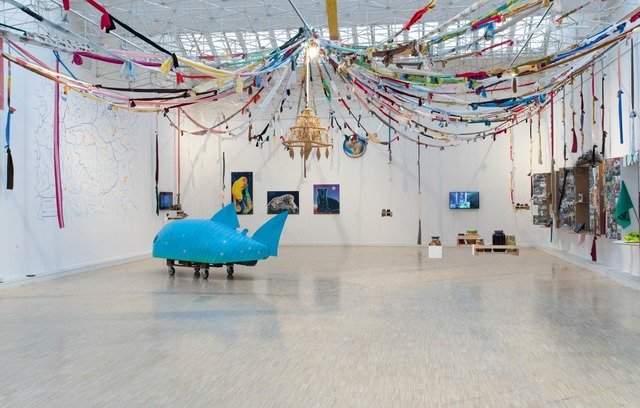 The picture presents the exhibition space shown from a wide perspective. There is a wooden floor and white walls. On the walls there are pictures and other exhibition objects, on the ceiling there is a metal structure. Colourful strips of fabric are suspended from it, spanning and extending from the centre of the ceiling to the walls. To the left is an object in the form of a blue fish on wheels made by artists duo: Cecylia Malik and Piotr Dziurdzia. Behind it are four paintings of animals, three in rectangular forms and one in an oval shape painted by Ewa Ciepielewska. 