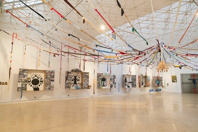 This is the same larger exhibition space shown from a different perspective to the previous photo. On the vault is a textile installation suspended from the centre to the walls. To the left is a series of wall-hung boxes resembling altars. They are dedicated to different themes like Earth, animals, forest, air and water. The altar-boxes contain black graphic signs depicting the aforementioned themes designed by artist Małgorzata Gurowska.