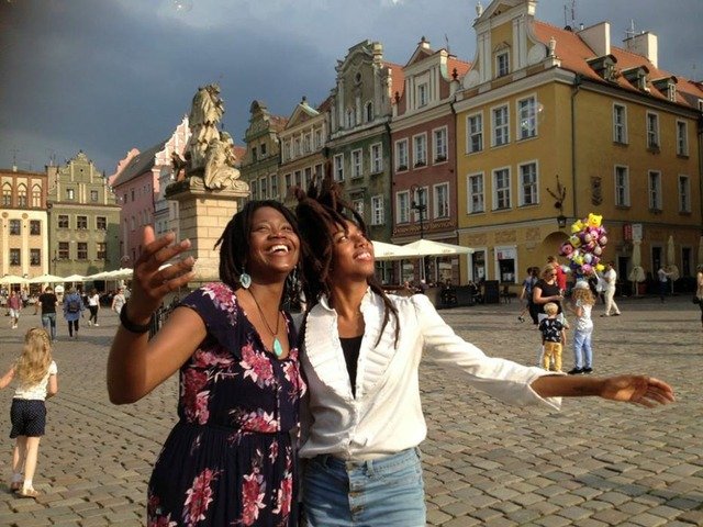 The photograph is a portrait of two Black, female-looking people, taken outside in the city's old town. It is a sunny day. The persons are smiling and pointing their hands upwards. Behind them is a statue and next to it stands a person selling colourful balloons.