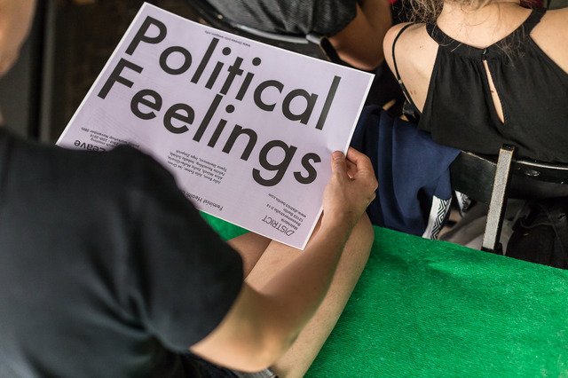 The image shows a person dressed in black holding a light purple poster with black letters: Political Feelings. The poster was made by the collective: Feminist Health Care Research Group.