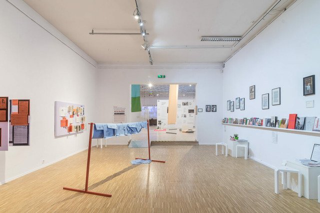 This is the first of six photos documenting the space of the exhibition “Creative Sick States: AIDS, CANCER, HIV” at the Municipal Gallery Arsenal in Poznan. The photo depicts a room with wooden floor and white walls. On the left is an installation of Inga Zimprich composed of leaflets and books hung on a light purple board. In front of the board is a red wooden rack on which blue paper brochures are hung. To the right is a bookshelf that runs the full width of the wall. Under the shelf are two white cubicle tables with three chairs each. Above the shelf, framed photographs hang on the wall. From this room one can get a view of the second, larger exhibition space.