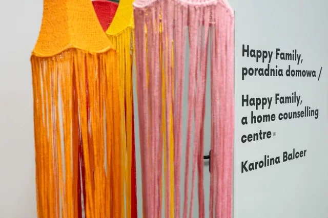 The first of six photos documenting the exhibit of Karolina Balcer “Happy Family, a home counselling centre” at the Municipal Gallery Arsenal in Poznan. It shows four crocheted lampshades in orange, pink, yellow and red. Behind them on the wall is an entry with the exhibition title in Polish and English.