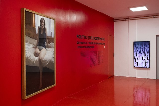 The first of the six photos documenting the exhibit “Politics of (In)Accessibilities, Citizens with Disabilities and Their Allies” at the Municipal Gallery Arsenal in Poznan. It shows a red painted wall and floor. On the left is a large-format photograph in wooden frames by Joanna Pawlik depicting a laughing girl with her right leg amputated above the knee. The girl is sitting on a bed, wearing only a T-shirt and panties. Next to the photograph is the exhibition title and an excerpt from the curatorial text. On the wall to the right is a television monitor with a video work by Daniel Kotowski.