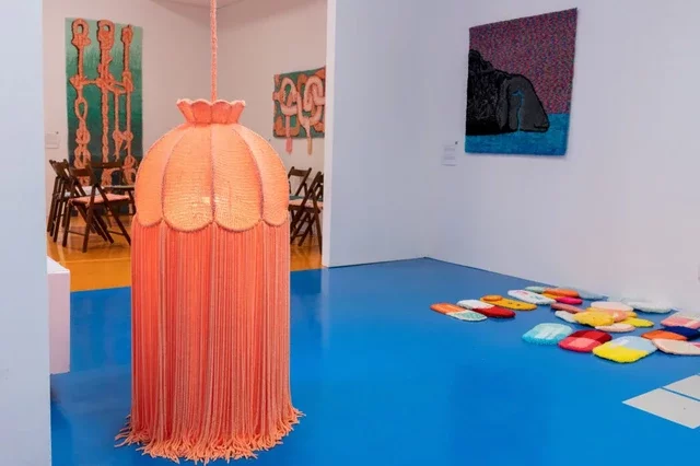 The second photograph is a view of the exhibition, with a large orange lampshade made by the artist in the foreground. Its tassels hang down to the blue-painted floor. Behind it on the left are wooden chairs arranged in a circle and two carpet paintings made by Karolina Balcer. To the right is another carpet painting depicting a black, crying sheep. Below it, hand-made colourful pills lie on the floor.
