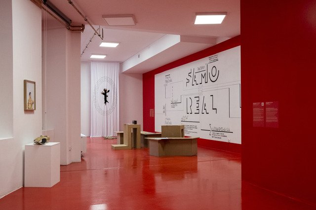 The view of the exhibition shows the space from a wide perspective. The floor is painted red, as is the wall on the right. The opposite wall is left white. On the right is an installation by Karolina Wiktor consisting of wooden panel objects and a wall painting with letters and signs. In front is Liliana Zeic's installation of fabric hanging from the ceiling to the floor. To the left is Pamela Bożek's work of portrait photography and a gold plaster removed from her son's leg.