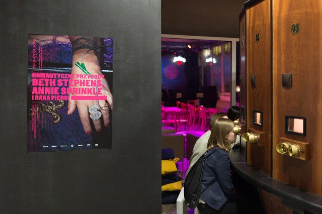 The second photograph shows part of the keiserpanorama and three people using it. On the left is an exhibition poster based on a photograph of Annie Sprinkle's hand with a silver ring with a dollar sign and a venflon inserted into one of the veins on her hand. The title of the exhibition is written in pink bold letters.