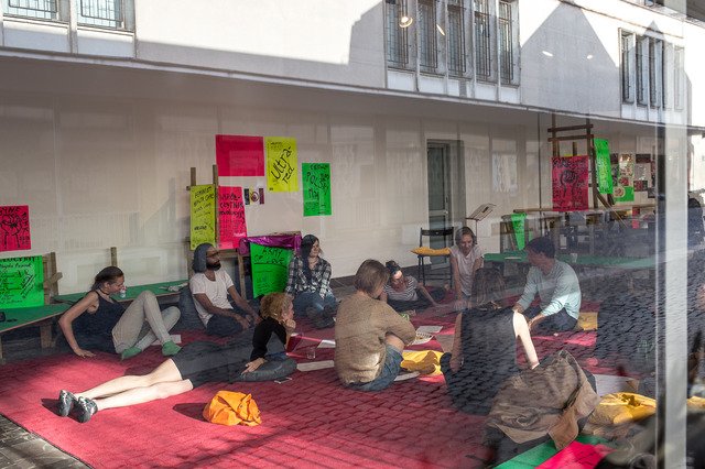 The second photograph documents the workshop of the Army of Love collective that took place in the exhibition space. A dozen people sit and lie on the red carpet. Around them hang colourful posters on recycled wooden constructions.