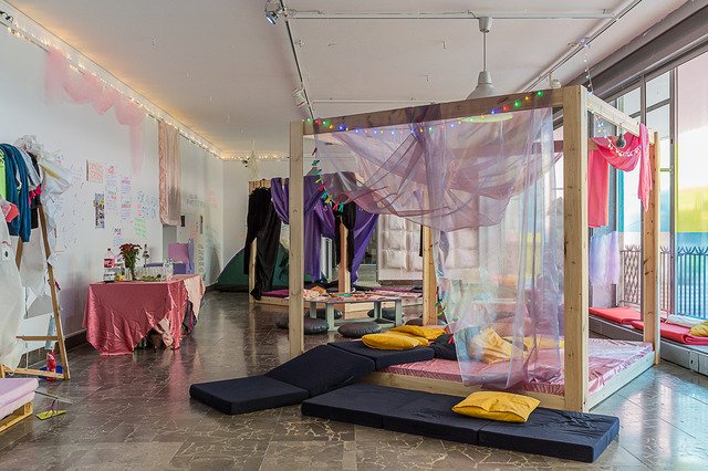The image shows the exhibition space from a wide perspective. In front, two dark blue mattresses lie on the floor. One of them is partly laid on a huge canopy bed, the other same bed stands just behind it filling almost the entire gallery space. Small Christmas tree lights hang from the ceiling. To the left stands a table with a bright pink tablecloth. To the right one can notice the windows, which are also the facade of the gallery. They are decorated with transparent foil in pastel colours.