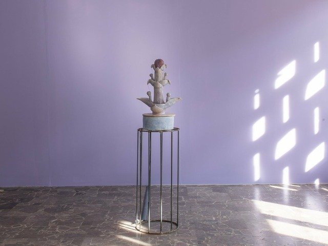 This is a photograph of an installation by Natalia Biało. It depicts a ceramic sculpture in the shape of a plant, with water flowing from it like a fountain. The sculpture is in light blue, purple and yellowish colours. It stands on a dark oval metal structure. From the sculpture comes a cable laid on the marble floor. In the background is a purple wall. The sun shines from the right illuminating the artwork.