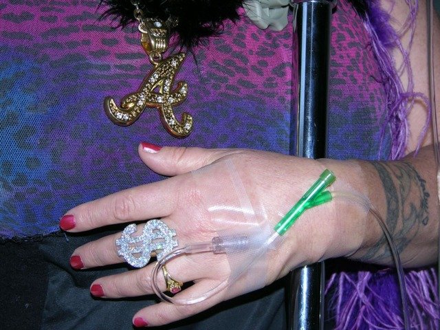 The photograph shows the hand of Annie Sprinkle with a silver ring with a dollar sign and a transparent green venflon tube inserted into one of the veins on her hand. The figure has her nails painted red. She is wearing a necklace with a gold A, and  a purple dress with a tiger pattern. There is a dark blue tattoo around her wrist.