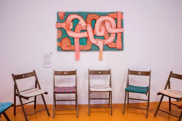 This image depicts wooden chairs with cushions on them. On the cushions are words in Polish embroidered by the artist that describe people in crisis in a negative, derogatory way: mad, crazy, hysteric, junky, psycho, etc. Behind the chairs is a carpet painting depicting joined bright pink hands and arms against a green background. The title of this work is 'Codependency'.