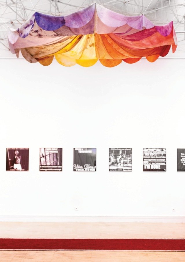 a photograph documenting an exhibition in the gallery, with graphic prints hanging on the wall and a colourful umbrella-like installation on the ceilings.