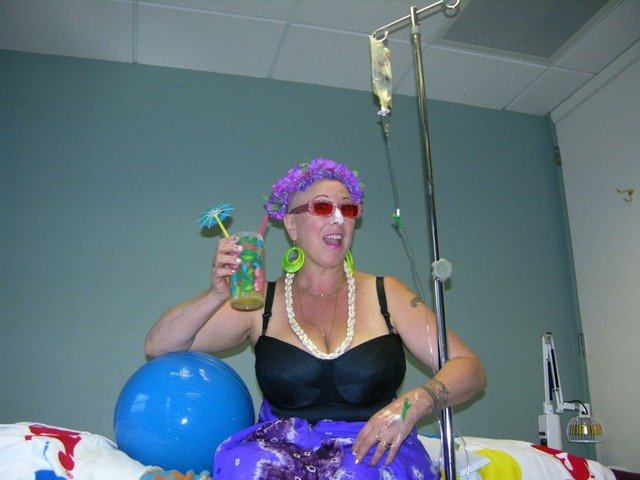 The photograph taken by Beth Stephens shows Annie Sprinkle on a hospital bed. She looks very cheerful, drinking a drink from a glass with a paper umbrella. She is wearing a black swimsuit and a shell necklace. On her head is a purple garland. A drip is attached to her left hand.  