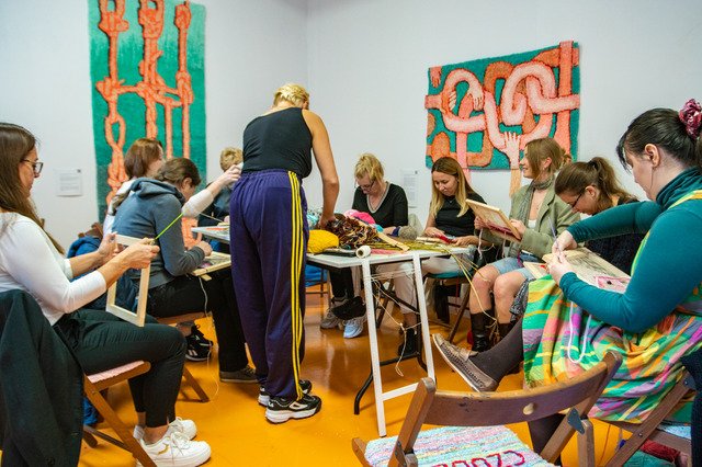The photograph shows a textile workshop led by Monika Winczyk and the Analog Group in the exhibition space. The people in the photograph are busy weaving. They are sitting on wooden chairs, a table with yarn and other materials stands in the middle. Behind them are two carpet paintings by Karolina Balcer.  