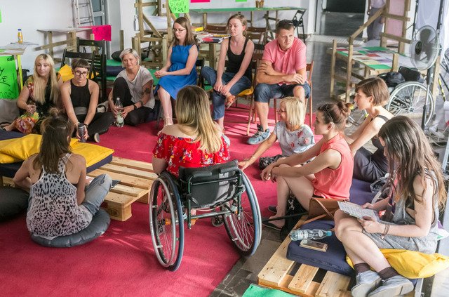 The last photograph documents a workshop run by a group of people with and without disabilities. Several people are sitting on a red carpet, on chairs or in wheelchairs. They are wearing summer clothes. It is a sunny day. The sun illuminates the exhibition space in the gallery. Behind the people are colourful posters hung on a wooden construction.