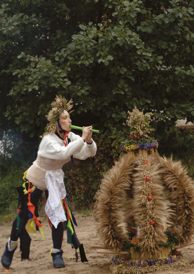 photograph depicting a person playing a flute, dressed in an extravagant variation on a folk costume. Next to it is an object made of hay.