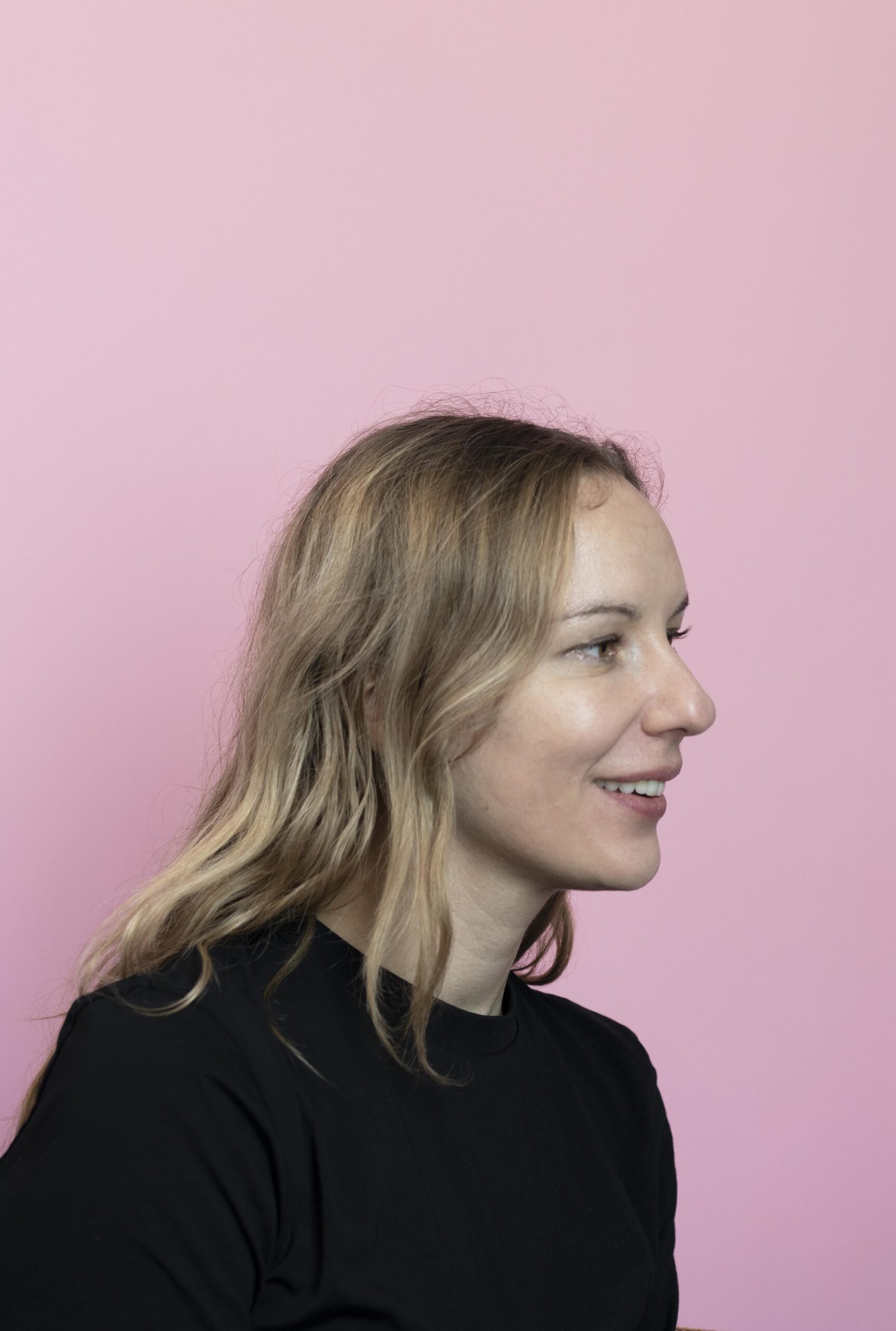 This is a portrait photo of Zofia nierodzińska, who is a female person, shown from her shoulders to the top of her head. She is wearing a black t-shirt. She has blonde, long hair. She is in her early thirties. The background is in a plain light pink colour.